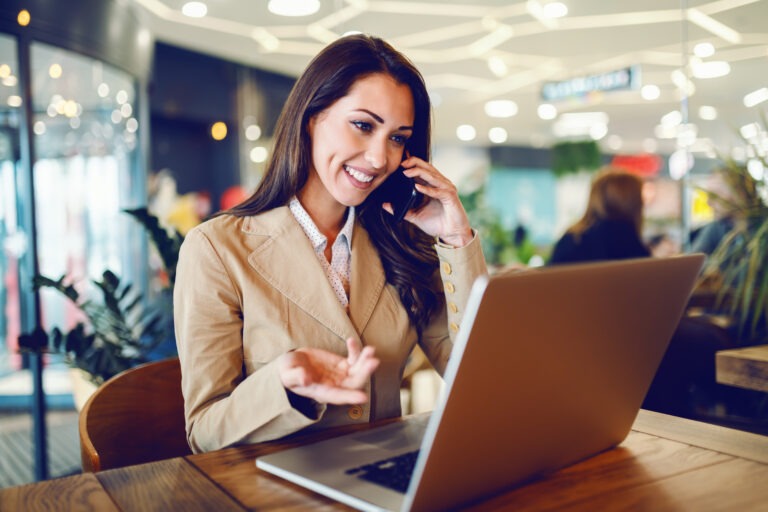 Attractive businesswoman in beige jacket sitting in cafe, talking on the phone and looking at laptop.